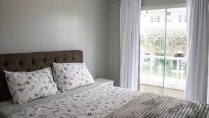 Un dormitorio con cabecera tapizada, ropa de cama floral y vistas a un balcón con cortinas blancas.
