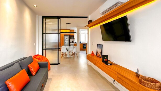 A modern living room with a grey sofa, orange cushions, a wooden console table, a flat-screen TV, and a kitchen in the background. - Timo Apto 350m da Praia de Copacabana RJ (Rio de Janeiro)