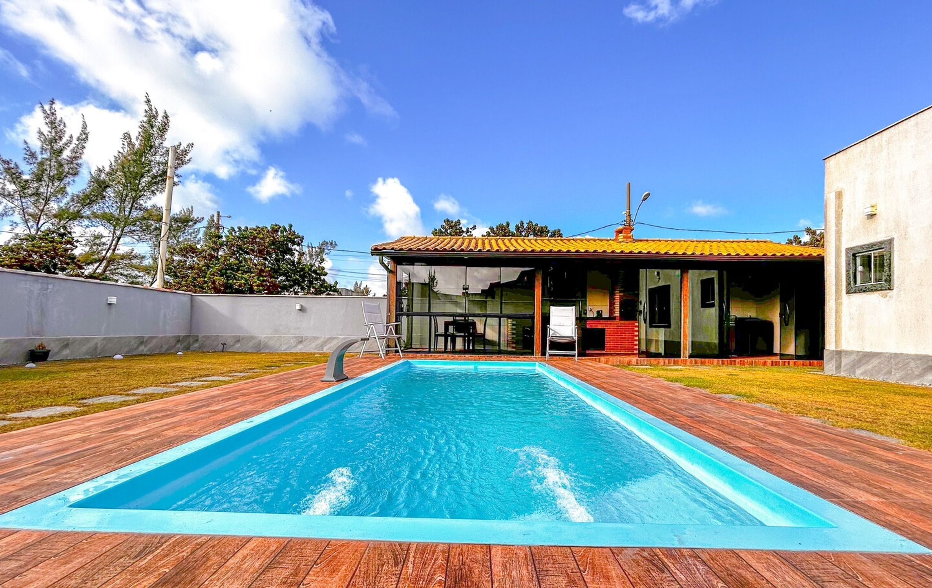 A rectangular swimming pool with a wooden deck, a covered patio area, and a white building with a tiled roof.