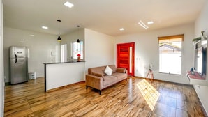 A modern living room with a sofa, a kitchen area, and a red door. - Casa a Poucos Metros da Praia do Coral - Cabo Frio (Cabo Frio)