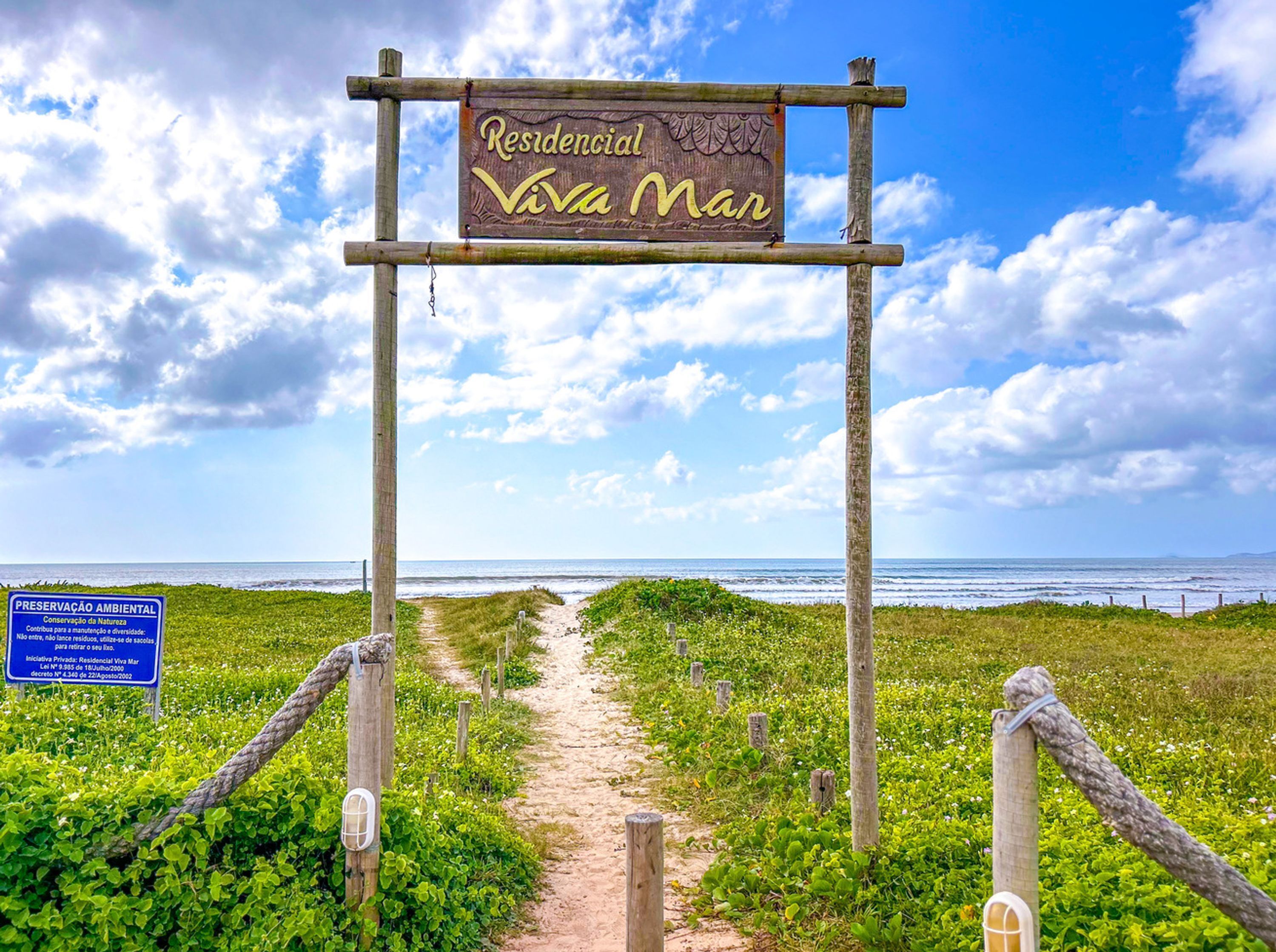 A wooden sign with "Residencial Viva Mar" written on it, leading to a beach path.