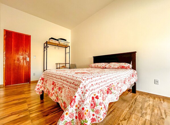 Une chambre avec un plancher en bois, un lit recouvert d’une couvre-lit à motifs floraux, une tête de lit en bois, une armoire et un portant à vêtements.