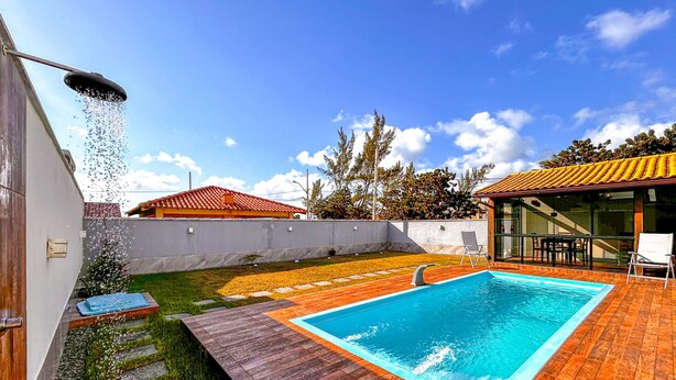 A swimming pool with a wooden deck, a covered patio area, and a view of a residential building with a red-tiled roof. - Casa a Poucos Metros da Praia do Coral - Cabo Frio (Cabo Frio)