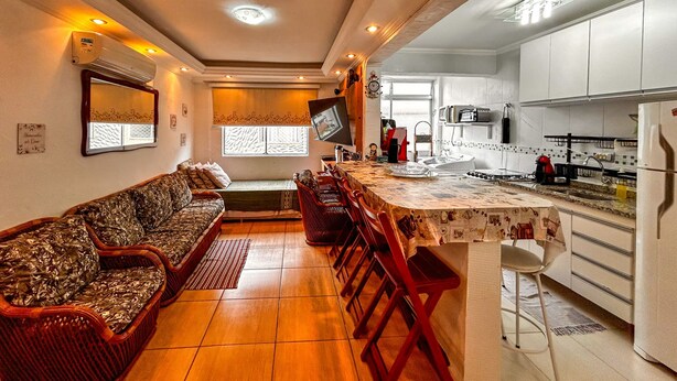 A cozy living room with a sofa and chairs, a kitchen with appliances, and a TV on the wall. - Apto a 800m da Praia da Enseada no Guaruj SP (Guarujá)
