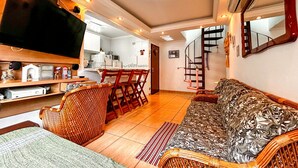 A living room with a sofa, a television, a kitchen area, and a staircase. - Apto a 800m da Praia da Enseada no Guaruj SP (Guarujá)