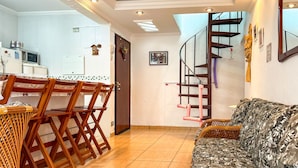 A living room with a sofa, a dining table with chairs, a kitchen area with a refrigerator, and a spiral staircase. - Apto a 800m da Praia da Enseada no Guaruj SP (Guarujá)