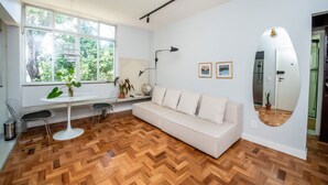 A modern living room with a white sofa, a round table, and two chairs. There is a large window with a view of greenery outside. - Apto com Excelente Localiza o em Asa Sul DF (Brasília)