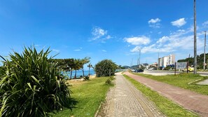 A paved pathway alongside a beach with a clear view of the ocean, palm trees, and a road with vehicles. - Casa Confort vel bem Equipada com Wi-fi e Garagem (Caraguatatuba)
