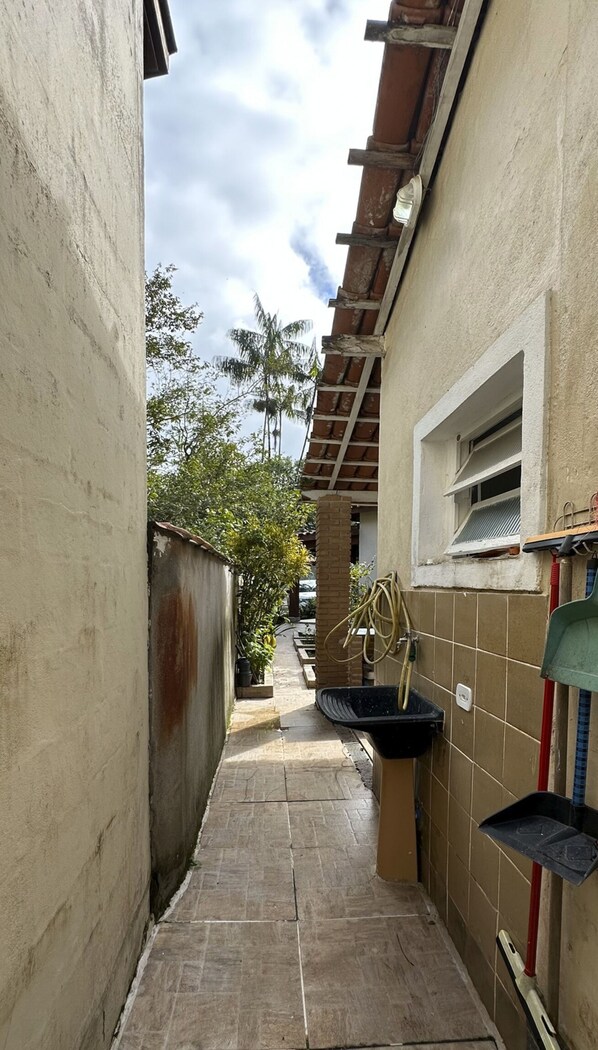 Une ruelle étroite au sol carrelé, une fenêtre ornée d’une grille métallique et un tuyau d’arrosage enroulé contre le mur.