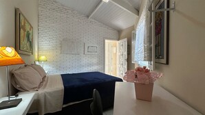 Ett hotellrum med en säng, ett skrivbord med en lampa, en stol, en vas med blommor och en vit tegelvägg.