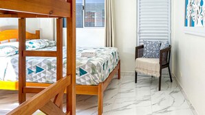 Ένα δωμάτιο με ένα διπλό κρεβάτι, ένα μονό κρεβάτι, μια καρέκλα, ένα παράθυρο με κουρτίνες και μια πόρτα με περσίδες.