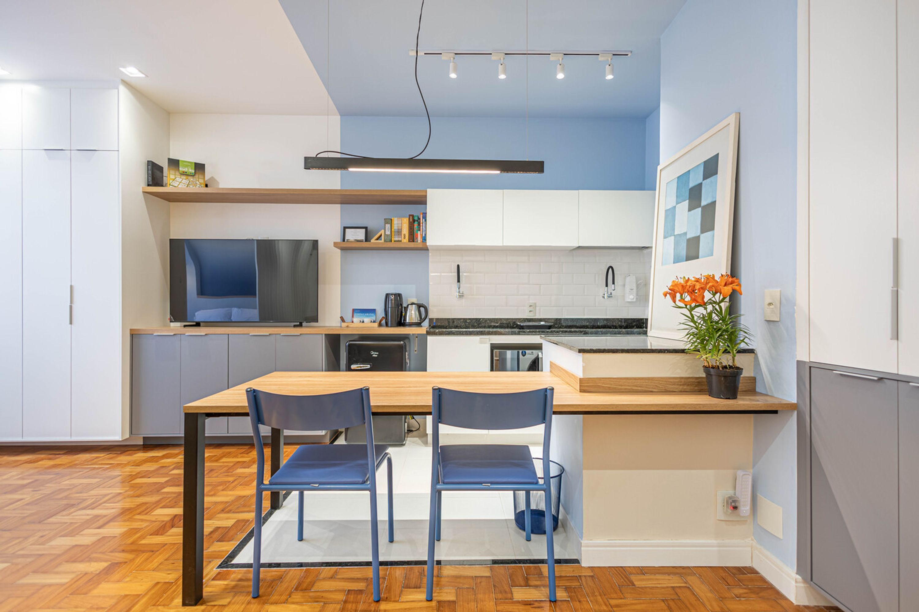 A modern kitchen with a wooden table, two blue chairs, a built-in oven, and a potted plant.