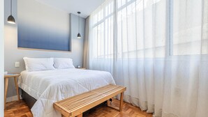 Une chambre à coucher avec un lit, un banc en bois et de grandes fenêtres ornées de rideaux légers.