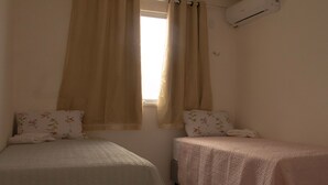 A bedroom with two beds, beige curtains, floral pillows, and a wall-mounted air conditioner.