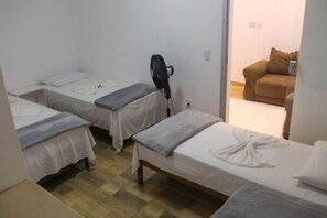 A room with three single beds, each with a white mattress and grey bedding. There is a black fan on one of the beds. The room has a wooden floor and a white wall. - Casa com Tima Localiza o em Porto Seguro BA (Porto Seguro)