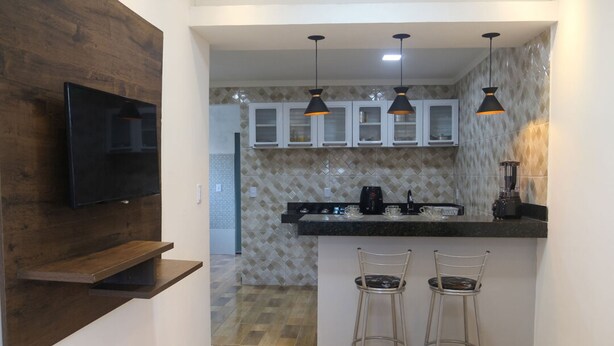 A modern kitchen with a wooden accent wall, a flat-screen TV, a black countertop, two bar stools, and pendant lighting.