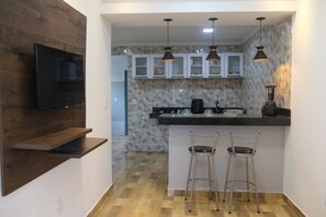 A modern kitchen with a wooden accent wall, a flat-screen TV, a black countertop, two bar stools, and pendant lighting. - Casa com Tima Localiza o em Porto Seguro BA (Porto Seguro)