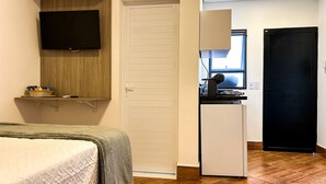 Une chambre avec un lit, un téléviseur fixé sur une étagère en bois, une petite kitchenette équipée d’un réfrigérateur et une porte donnant sur une autre pièce.