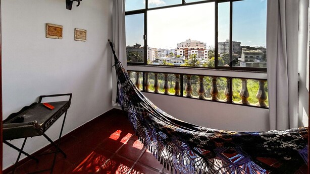 A room with a hammock, a small table, and a view of buildings through a large window. - Apto a 210m da Praia da Pontinha em Araruama RJ (Araruama)