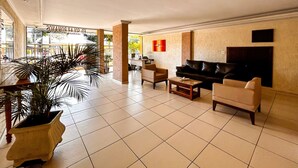 A spacious lobby with a large potted plant, a leather sofa, a coffee table, and a reception desk. - Apto a 210m da Praia da Pontinha em Araruama RJ (Araruama)