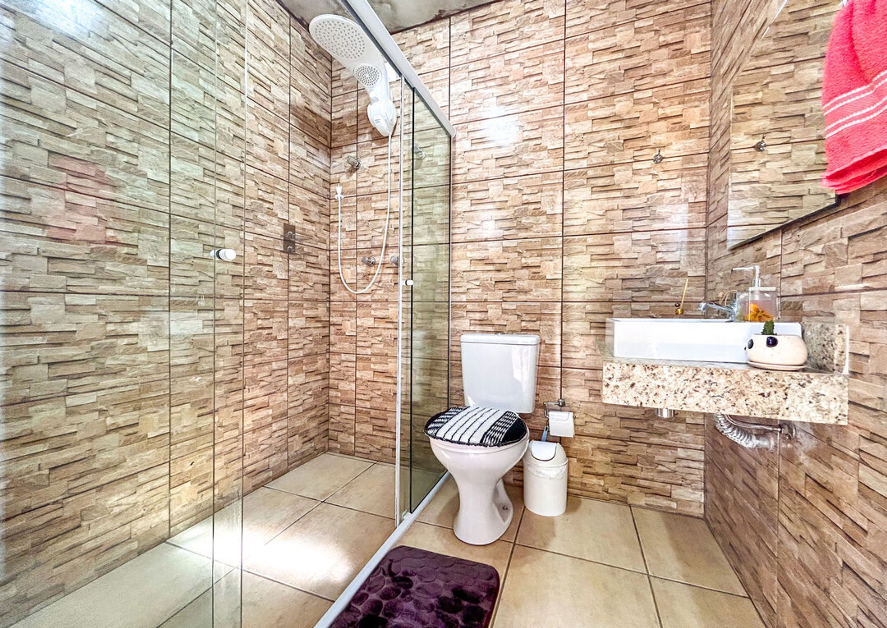 Un baño con ducha de vidrio, inodoro, lavamanos con encimera de mármol y espejo con una toalla roja colgada.