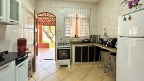 Cocina compacta con muebles blancos, estufa, refrigerador y pileta. Se ve una ventana con cortinas de encaje y vistas a la vegetación.