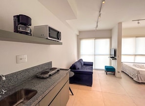 Cocina moderna con microondas, cafetera y horno empotrado. En la sala de estar, hay un sofá y una cama, junto con un televisor sobre un mueble.
