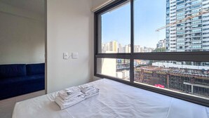A hotel room with a large window, a bed with white linens, a blue sofa, and a wall-mounted light switch. - Timo Apto a 900m do Parque Ibirapuera com Wifi SP (São Paulo)