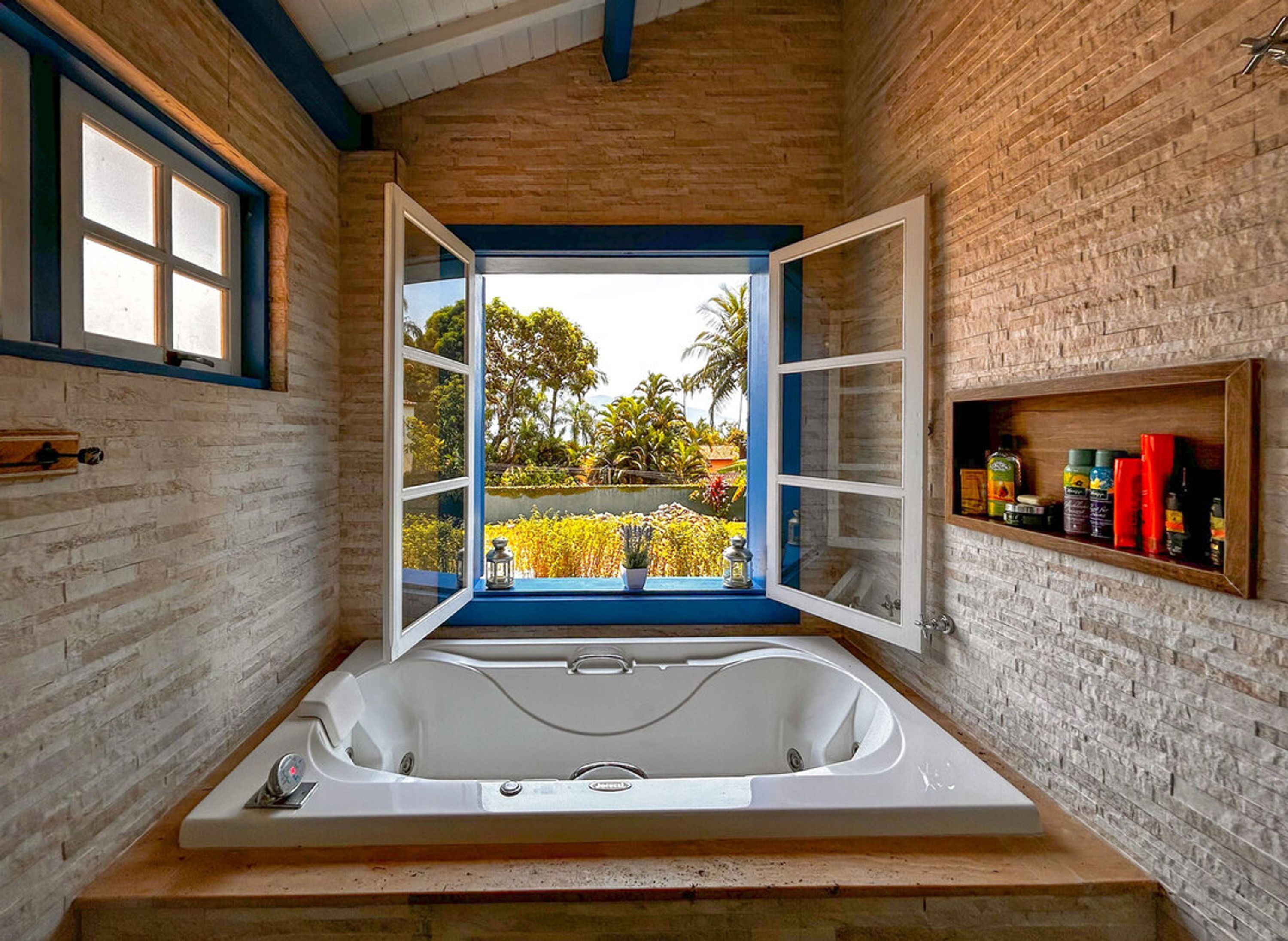 A bathroom with a large bathtub, a window with open shutters, and a shelf with toiletries.