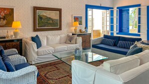 A living room with white brick walls, blue shutters, and a glass coffee table. - Casa pr x Praia de Santa Teresa Ilhabela SP (Ilhabela)