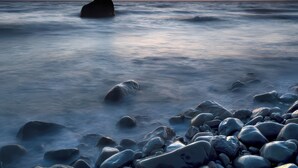 A rocky shoreline with a large rock formation in the distance during sunset.
