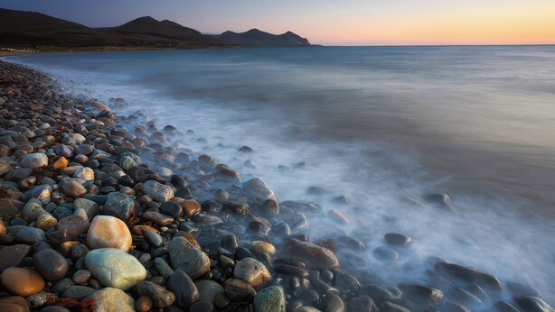A pebbled beach with a calm sea and distant mountains.
