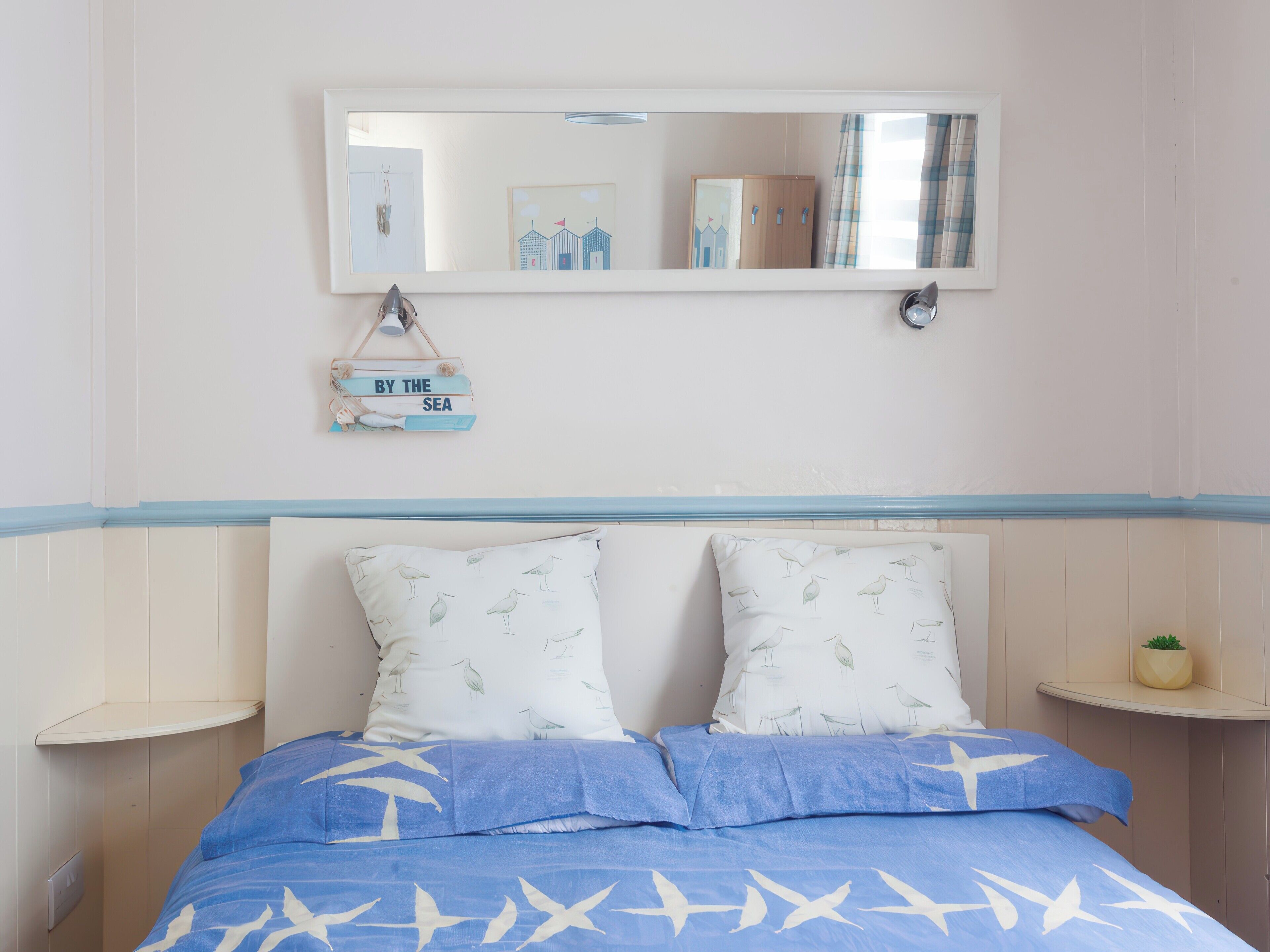Un dormitorio con cama, dos almohadas, una mesita de noche, un espejo, un estante en la pared y una planta pequeña.