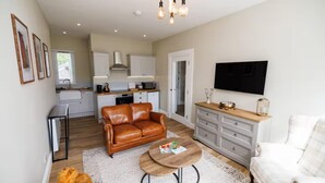 Una sala de estar acogedora con un sofá de cuero marrón, un sillón blanco y una mesa de centro de madera. En la pared hay un televisor, y se aprecia una cocina al fondo.