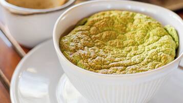 A white bowl of green dessert on a white plate with a saucer, accompanied by a small bowl of bread and a white cup in the background.