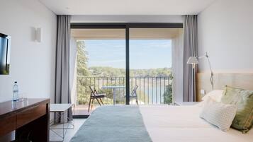 Una habitación de hotel moderna con una cama grande, un escritorio, una silla y un balcón con vistas a un lago y árboles.