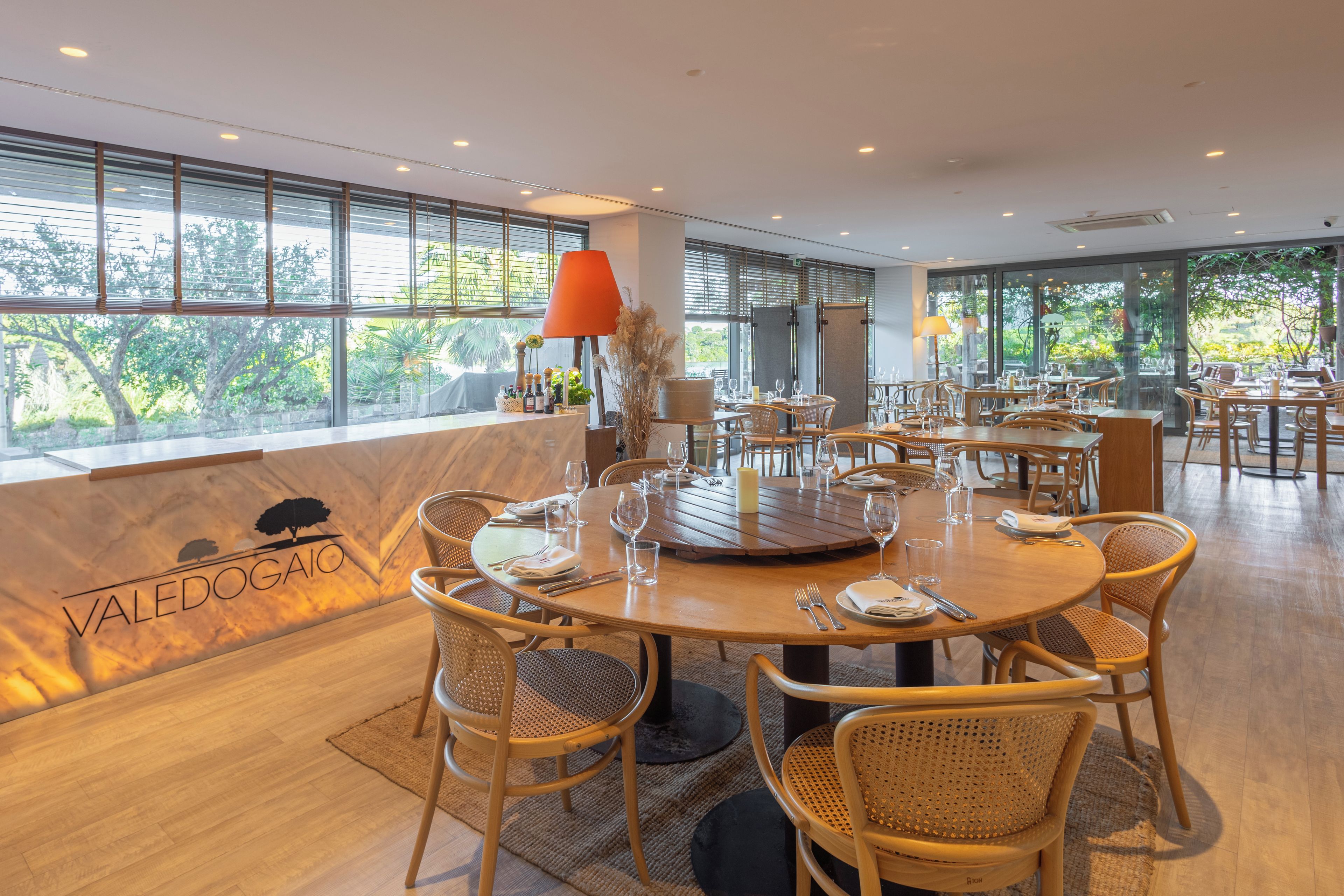 A restaurant with round tables and wicker chairs, large windows with a view of trees, and a wooden floor.