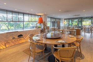 A restaurant with round tables and wicker chairs, large windows with a view of trees, and a wooden floor. - Hotel Vale Do Gaio (Alcacer do Sal)