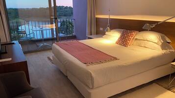 A hotel room with a large bed, a desk, a chair, and a balcony with a view of a body of water and greenery.