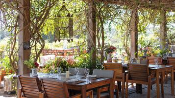 An outdoor dining area with wooden tables and chairs, surrounded by greenery and a pergola structure.