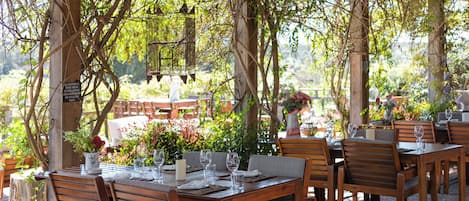 An outdoor dining area with wooden tables and chairs, surrounded by greenery and a pergola structure.