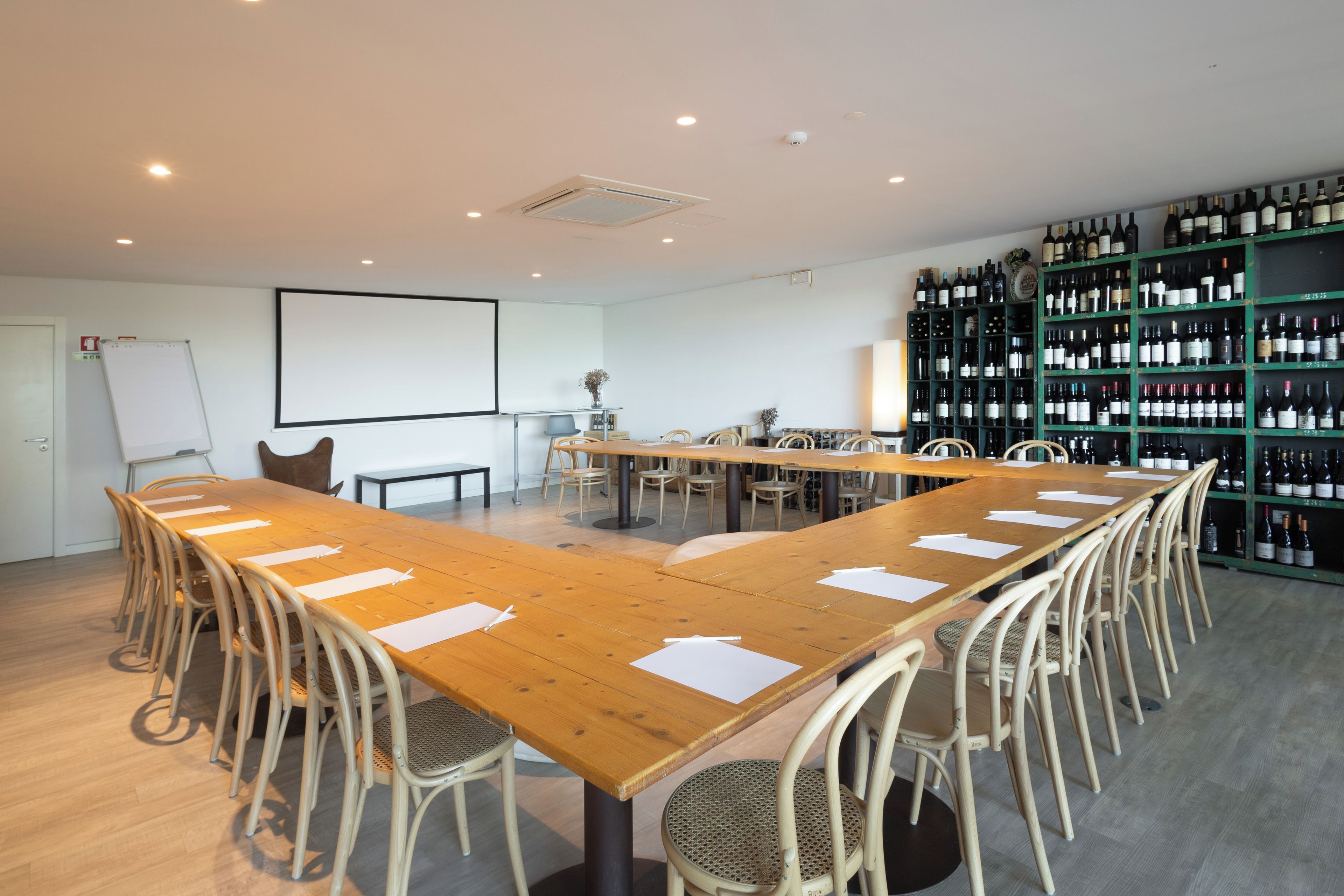 A conference room with a long wooden table, multiple chairs, a whiteboard, a projector screen, and a wine rack with numerous bottles.