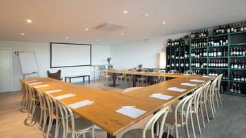 A conference room with a long wooden table, multiple chairs, a whiteboard, a projector screen, and a wine rack with numerous bottles.