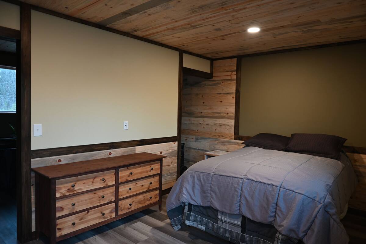 Une chambre avec une tête de lit en bois, un lit recouvert d’une couverture grise, une commode en bois et un mur d’accent en pierre.