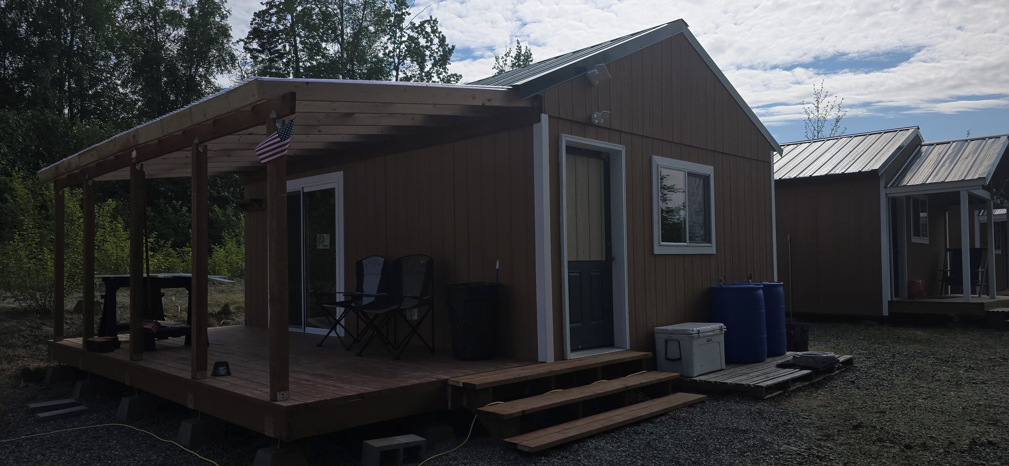 A wooden cabin with a porch, a small table, two chairs, and a trash bin.