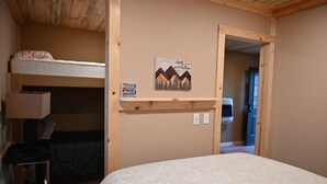 A bunk bed room with a wooden ceiling, a bed with a patterned bedspread, a small table, and a wall-mounted shelf with a picture and a sign. - Alaska River Pirates Cabins (Soldotna)