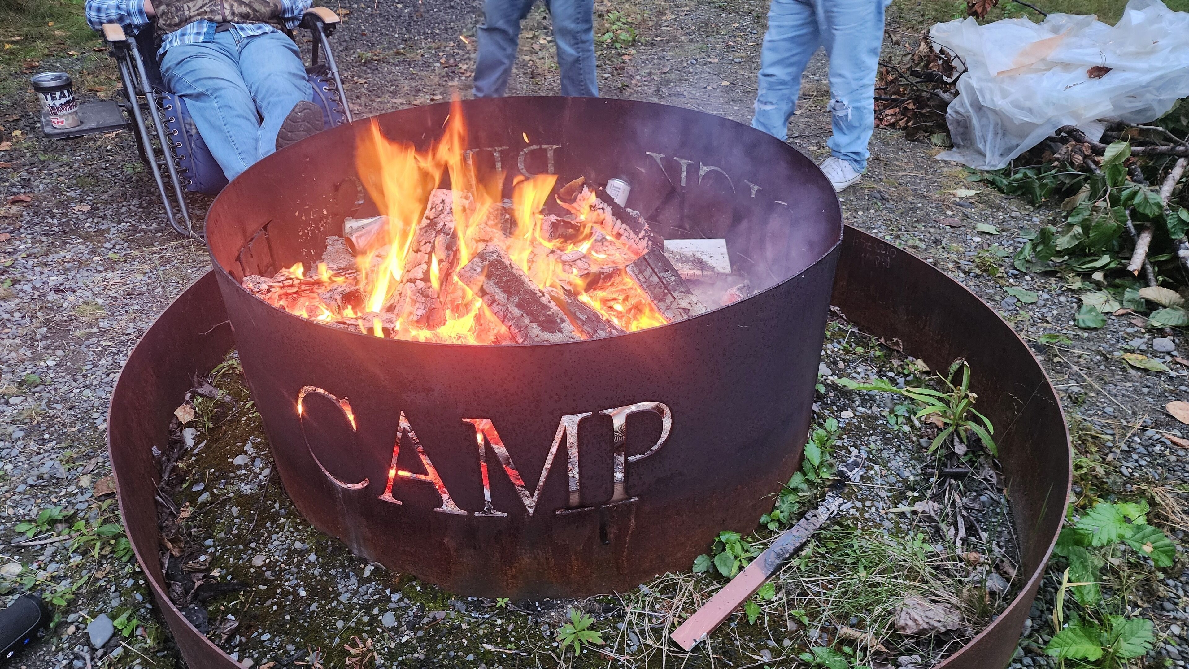 Een metalen vuurkuil met daarop het woord “CAMP”, omringd door grind en begroeiing.