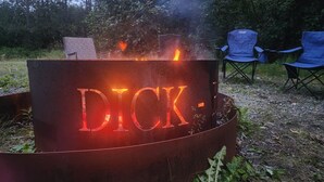 A metal fire pit with the word "DICK." engraved on it, surrounded by greenery and two blue chairs. - Alaska River Pirates Cabins (Soldotna)