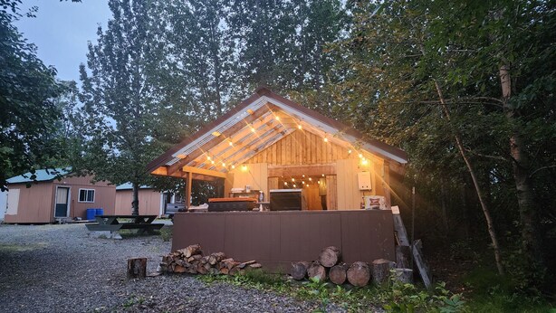 A rustic wooden structure with a sloped roof and string lights, surrounded by trees and a gravel path. - Alaska River Pirates Cabins (Soldotna)