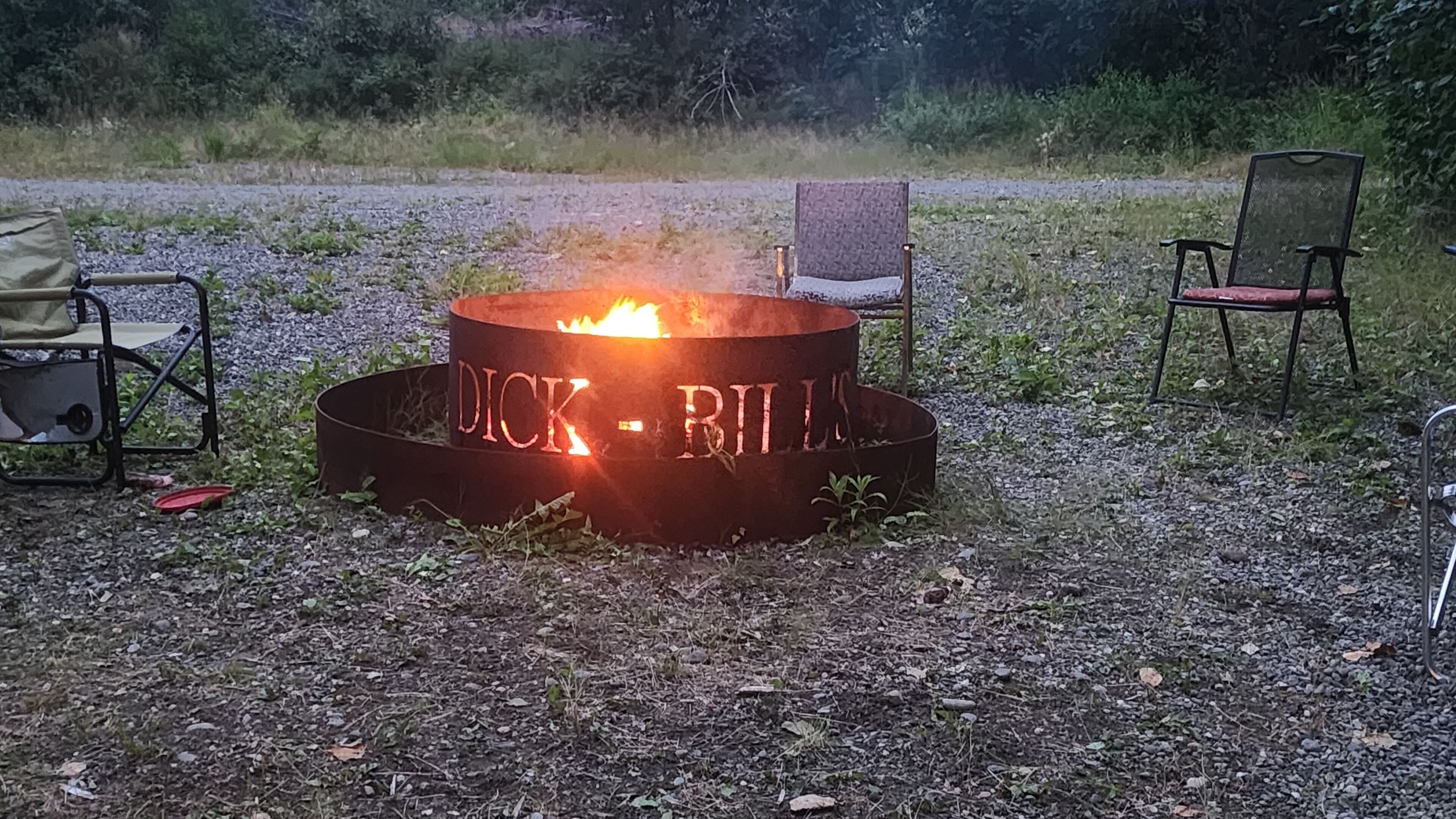 A metal fire pit with the inscription "DICK - BILL" surrounded by gravel and grass, with two chairs nearby.